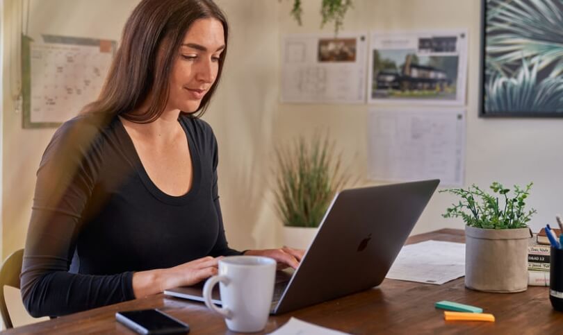 A builder uses her laptop on a desk with a mug and her phone nearby