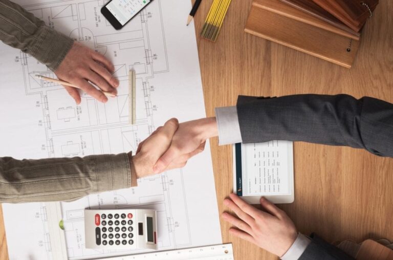 A bird's eye view of two people shaking hands with a tablet using Buildxact, paper plans, phone and calculator underneath