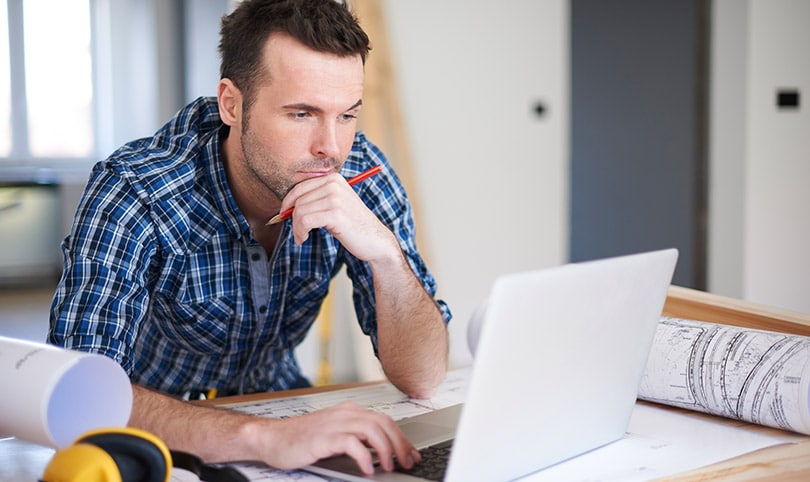 A builder working at a laptop surrounded by paper plans