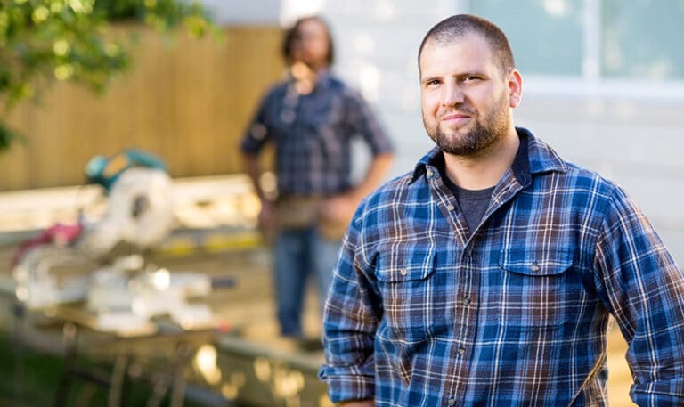 A builder on a job site facing the camera, with another builder on the tools in the background