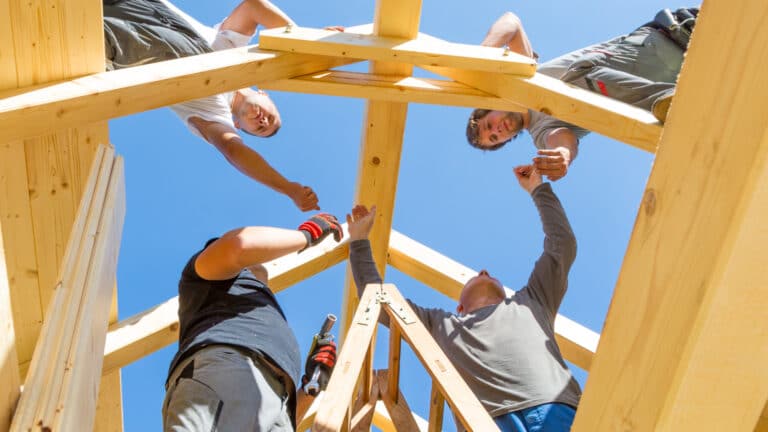 A team of builders work together on a rafter while constructing a roof of a home.