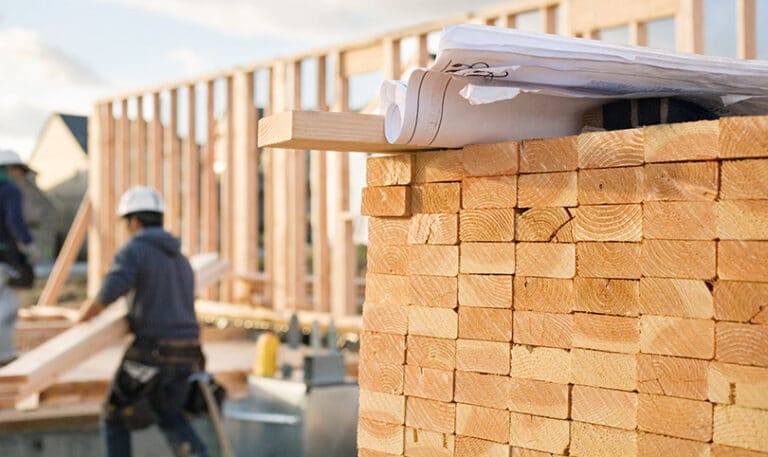 Plans laying on top of a pile of lumber on a construction site with a builder in the background