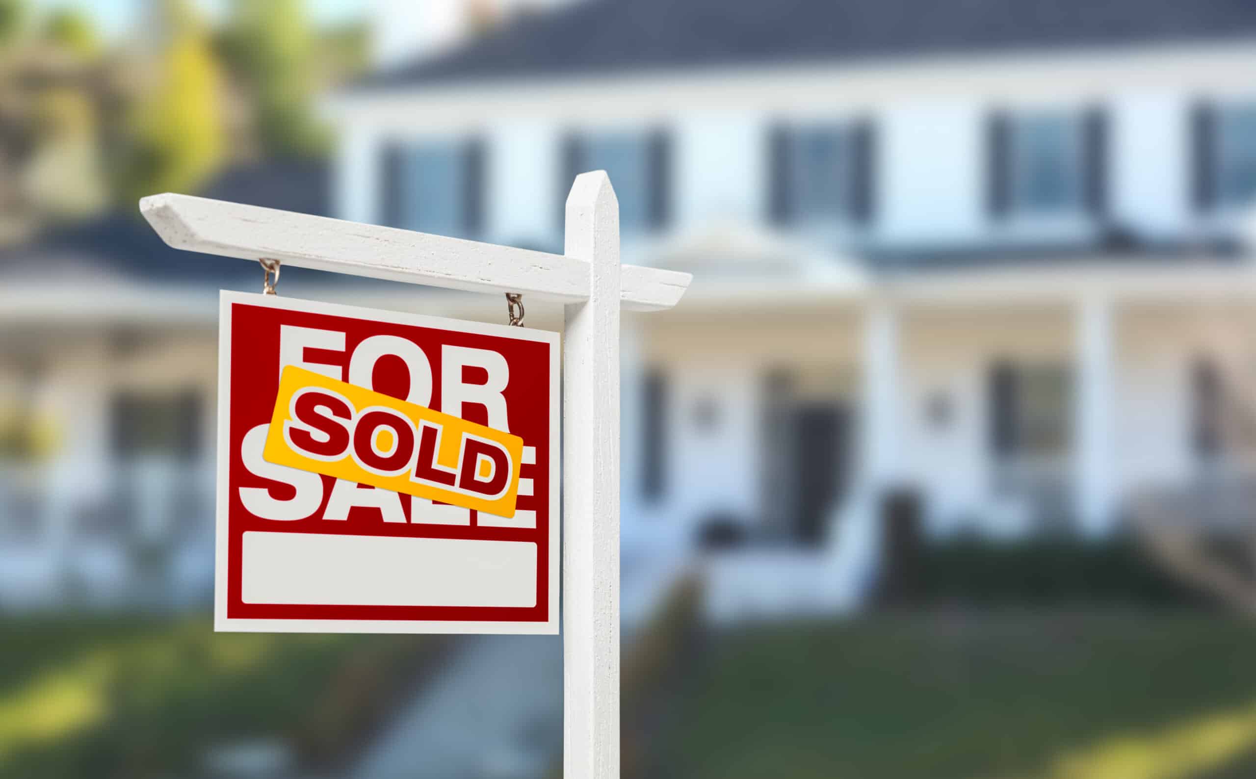 A 'For Sale' sign with a 'Sold' sticker placed on it in front of a large with house in the background
